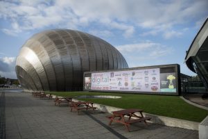 Image of Glagow Science Centre with large screen filled with logos of organisations involved in Discover Digital. Blue sky and green grass fame the picture.