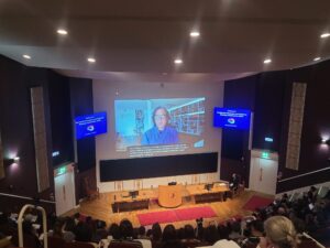 A large auditorium filled with people. A large screen at the front displays a video call with Professor David Olusoga who is wearing glasses and a blue shirt.