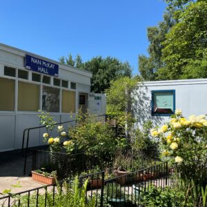 A white building with a blue sign above the entrance reading ‘Nan McKay Hall’. The entrance has a ramp leading up to an open door. The area is surrounded by lush greenery and blooming yellow roses, with another white building visible in the background. The sky is clear and blue.