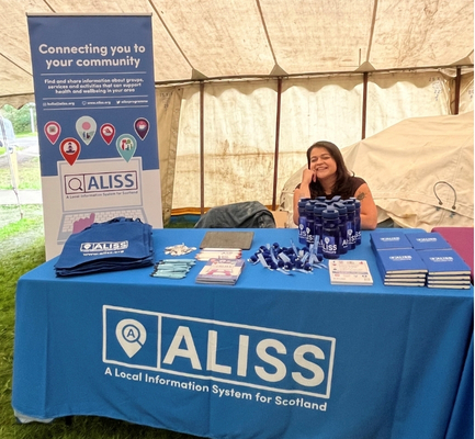 A woman with dark hair sitting behind a display table. The table is covered with a blue ALISS tablecloth and on top there are flyers, pens, tote bags, and water bottles. Behind the table is a blue banner with hands typing on a laptop and colourful location pins. The text on the banner reads: "Connecting you to your community".