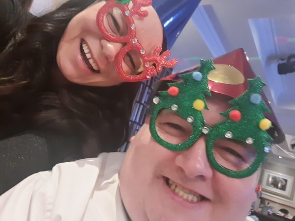 Photo of John smiling wearing novelty Christmas glasses in the shape of Christmas trees, with a woman, smiling, also wearing novelty Christmas glasses with antlers.