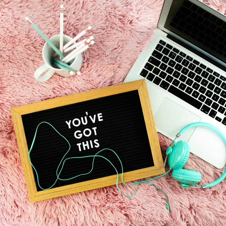 Birds eye shot of a laptop sitting on a carpet, next to headphones and a mug full of pencils. A framed sign with text reads "You've got this"