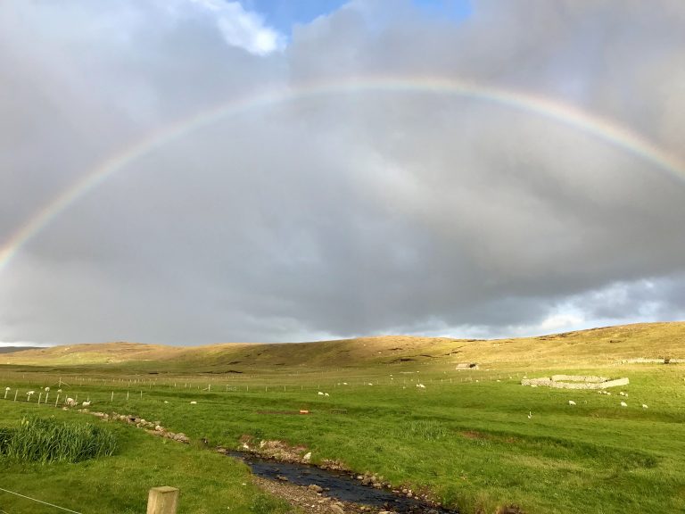 Picture of a field, with green grass and white sheep grazing. There is a bubbling blue brook in the foreground, with small light brown boulders edging the brook. There are small grass covered hills in the background which have shadows from the clouds blocking the sun on them. The clouds in the sky at the top of the image are grey and you can just see a hint of blue sky where the clouds break. There is a faint rainbow, at the front of the clouds reaching from the left of the picture to the right of the picture.