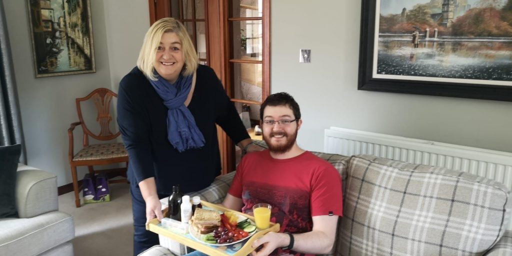 Young man sat on the sofa with food on a tray, smiling, with his mother stood next to him, smiling.