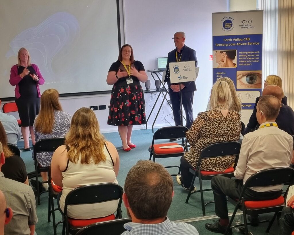 Image shows a photo of a room at Forth Valley Sensory Centre taken during the launch of the Forth Valley CAB Sensory Loss Advice Service on the 24th of July 2025. Presenters are standing at the front of the room beside a CAB banner advertising the new Service. Attendees are seated facing the presenters.