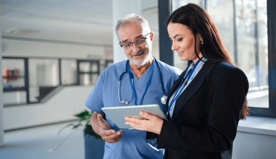 Image of an older male doctor in scrubs being shown a tablet screen by a training specialist