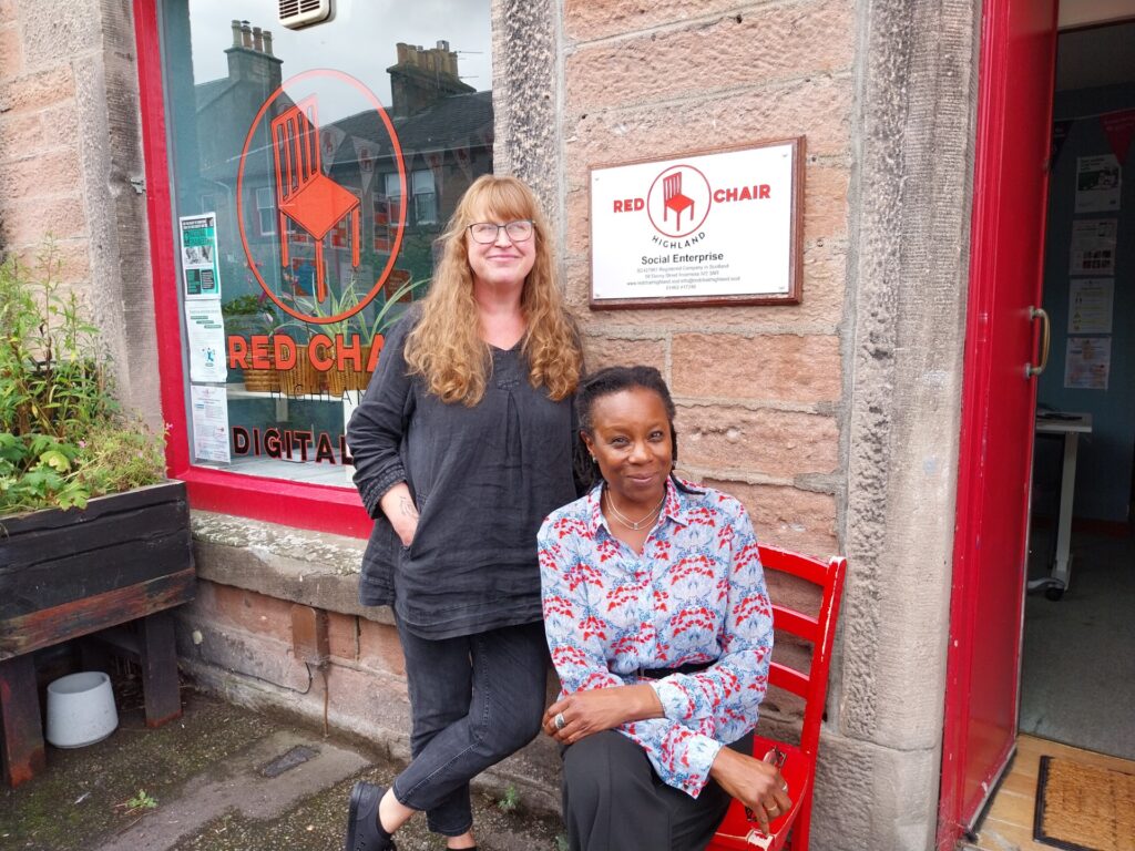 Two women smiling in front of a window and a sign with Red Chair Highland Social Enterprise on, in red.