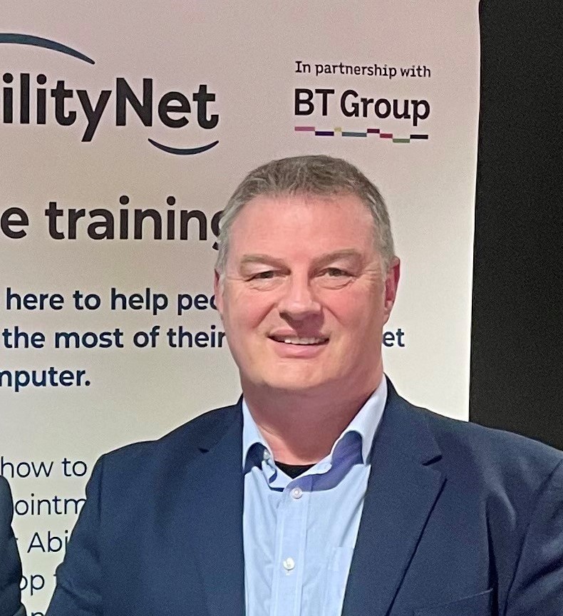 Rob, an older man in a suit smiling while at an event with AbilityNet banner behind him