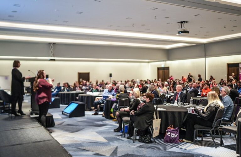 A photo from the annual alliance conference. The image shows a conference hall filled with people sitting at tables. All of the people are facing a stage on the left side listening to a speaker.
