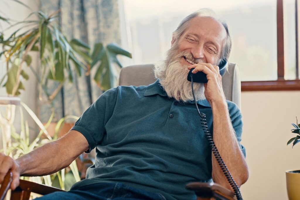 An older bearded man smiles while talking on a landline in a sunny room