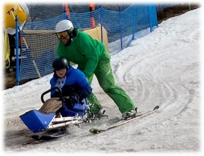 Darren sitting on sleigh being supported down snow slope by male instructor